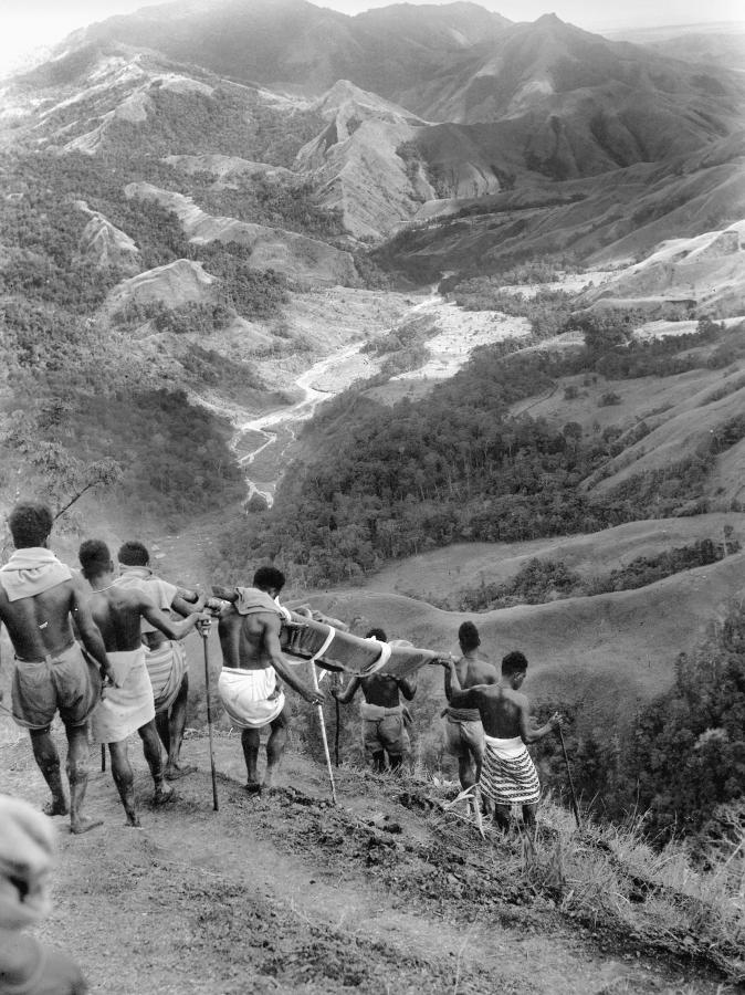 Native bearers carrying a stretcher case down a steep slope from Shaggy Ridge, on the way to the Advanced Dressing Station at Guy's Post. Centre is the Faria River, and in the background the Ramu Valley.