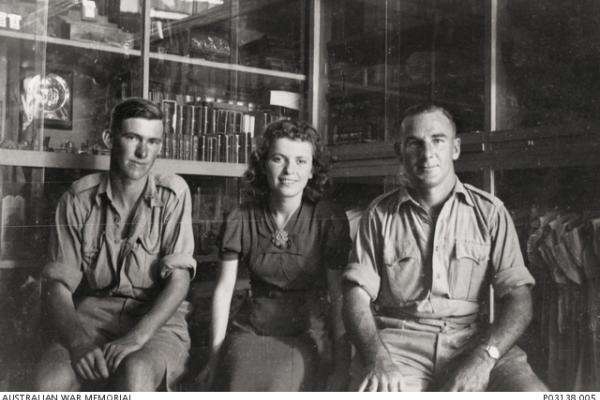 Informal portrait of VX4796 Private (Pte) Eric Clifton Eldridge, Headquarters, 17th Brigade, Australian Infantry, of Horsham, Vic (left); a shop girl (centre); and VX1113 Corporal (Cpl) Henry James Bailey Spencer (enlisted as Henry Bailey Spencer), 2/7th Battalion, of Alphington, Vic (right) inside a shop selling curios and childrens’ clothes in Tel Aviv. 